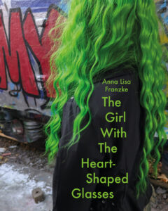 Eine grünhaarige Person steht vor einem Graffito. Auf ihrer schwarzen Jacke sind Autorin und Titel angegeben: Anna Lisa Franzke "The Girl With The Heart-Shaped Glasses".